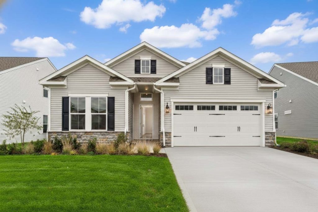A bright, daytime exterior of a new construction single-family home at Serenity Walk at Plainsboro, NJ, featuring a light gray facade, stone base accents, a two-car garage, and a manicured green lawn under a blue sky.